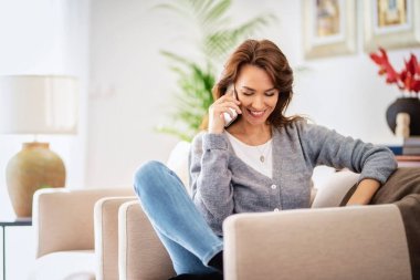 Çekici esmer saçlı kadın evde dinlenirken günlük kıyafetler giyiyor ve akıllı telefon kullanıyor.. 