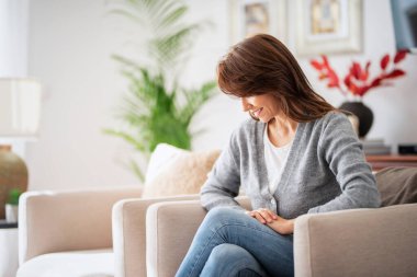 Evdeki koltukta dinlenen bir kadının fotoğrafı. Kumral kadın, kazak ve kot pantolon giyiyor ve düşünceli görünüyor..