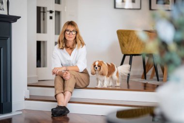 Orta yaşlı, mutlu bir kadın yerde beyaz şirin köpeğiyle oturuyor. Evde dinlenirken gözlük takan çekici bir kadın..