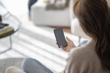 Koltukta dinlenirken elinde akıllı telefon tutan tanınmayan bir kadının arka plan görüntüsü. Boş ekran şablonu oluşturulsun. Omuz manzarasının üstünde..