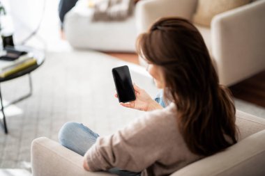 Koltukta dinlenirken elinde akıllı telefon tutan tanınmayan bir kadının arka plan görüntüsü. Boş ekran şablonu oluşturulsun. Omuz manzarasının üstünde..