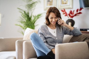 Çekici esmer saçlı kadın evde dinlenirken günlük kıyafetler giyiyor ve akıllı telefon kullanıyor.. 