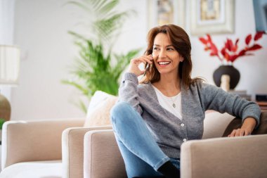 Çekici esmer saçlı kadın evde dinlenirken günlük kıyafetler giyiyor ve akıllı telefon kullanıyor.. 