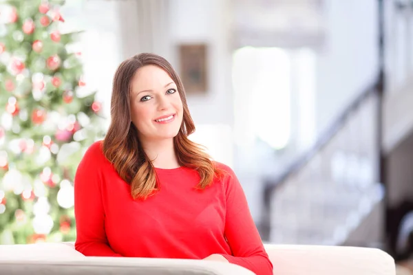 照片上的年轻女子坐在客厅的沙发上 微笑着看着相机 背景中的圣诞树 — 图库照片