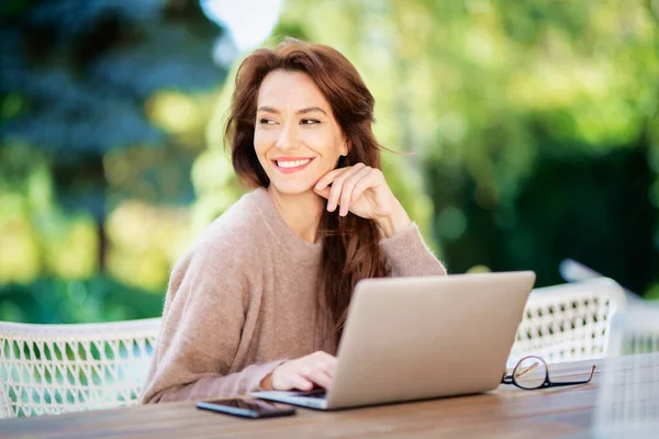 バルコニーに座ってノートパソコンを持つ自信を持って中年の女性 魅力的な女性はセーターを着て陽気な笑顔 — ストック写真