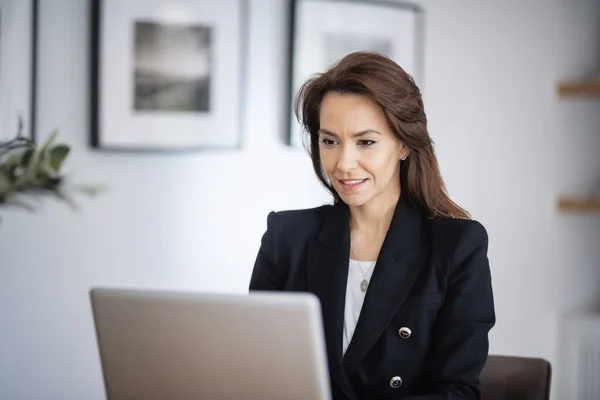 Shot of an attractive business woman using a laptop while working in her home office. Confident female wearing blazer.