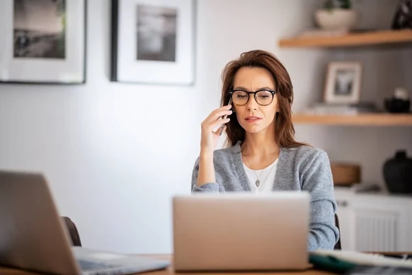 Thinking woman wearing eyewear and casual clothes while working from home. Business woman using laptops and making a call. Home office.