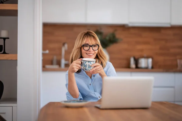 用笔记本电脑微笑中年妇女 在家里工作的时候有视频通话 自信的女性坐在厨房的餐桌边喝茶 — 图库照片