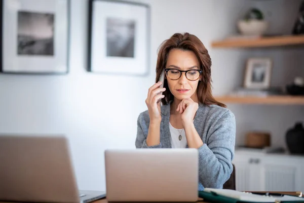 Thinking woman wearing eyewear and casual clothes while working from home. Business woman using laptops and making a call. Home office.