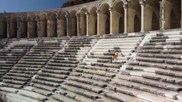 Leuke vrouw toerist in gele jurk wandelen in het oude amfitheater Aspendos — Stockvideo