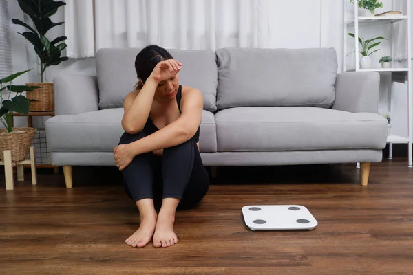 Young Asian woman sitting on the floor looking at the weight scale worried and depressed in dieting and eating disorder concept