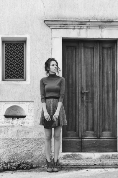 ブーツを着た美しいスレンダーな流行の若い女性のモノクロームの肖像画と通りで待っている古い木製のドアの前に立つミニスカート — ストック写真