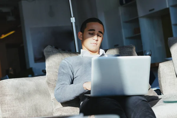 仕事に集中しながら暖かい日差しの中で家に座っている快適なソファの上でノートパソコンで作業している黒人 — ストック写真