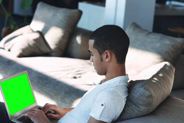 若い男リラックスしているソファの上で自宅から働いている間にCovid 19パンデミック彼の空白の緑のラップトップ画面の肩のビューで — ストック写真