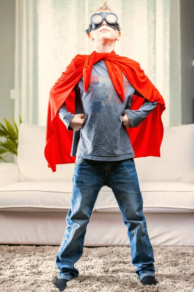 Proud young Super Hero posing in a red cape and goggles with hands on hips in the living room at home