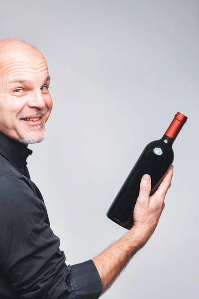 一位快乐的老人拿着一整瓶红酒 转身对着摄像机笑着 因为他期待着能在近距离拍摄的场景中喝到红酒 — 图库照片