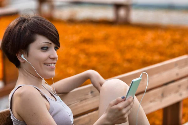 时尚的年轻女子坐在长椅上 在近距离观看音乐时 带着幸福的微笑听着音乐 — 图库照片