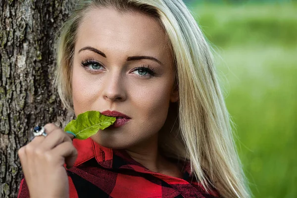 年轻美丽的金发女子靠在一棵树上 在草地或树林中央 把一片叶子放在嘴唇上 — 图库照片