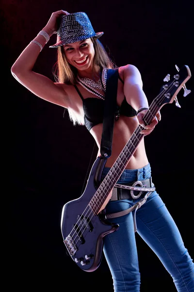 Fun loving vivacious young female guitarist smiling at camera with a beaming smile and hand to her sequinned hat as she stands on stage with her electric guitar