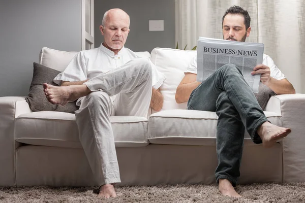 家で快適なソファに座っている間 印刷された新聞からビジネスニュースを読んでいる父と息子 — ストック写真