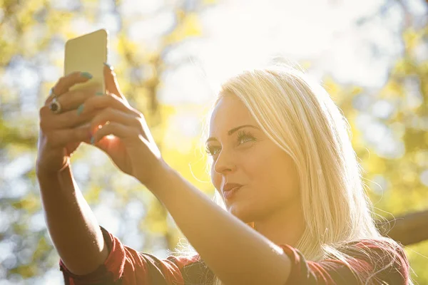 木の後ろに太陽の暖かい輝きに照らされた携帯電話でセルフィーを撮る若い女性の高いキーイメージ — ストック写真