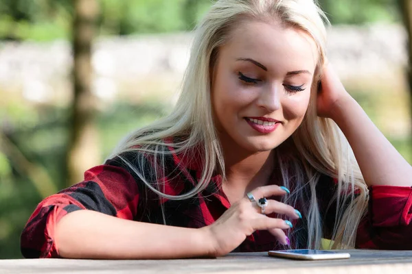 魅力的なトレンディーなブロンド女性タイプAテキストメッセージ上の彼女の携帯電話上の彼女は屋外のテーブルに座っている — ストック写真