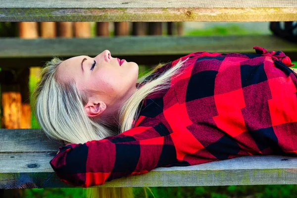 在公园的长椅上睡觉或打盹的安详的年轻女子 手放在后脑勺上 身穿五颜六色的红色衬衫 近视眼 — 图库照片