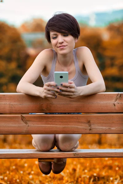 彼女は静かな笑顔で背中にもたれるように彼女の携帯電話を使用して公園や庭で素朴な木製のベンチにひざまずいて若い女性 — ストック写真