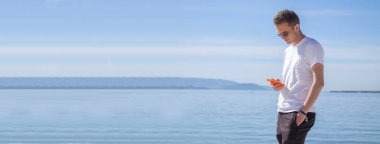 Genç adam sahilde yürüyor ve cep telefonu kullanıyor.