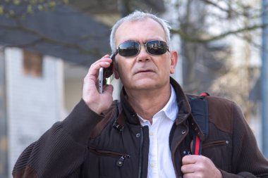 Şık, olgun bir adam sırt çantası ya da çantasıyla şehir kaldırımında yürüyor ve cep telefonunu kullanıyor.