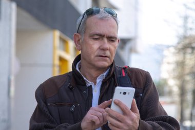 Şık, olgun bir adam sırt çantası ya da çantasıyla şehir kaldırımında yürüyor ve cep telefonunu kullanıyor.