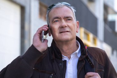 Şık, olgun bir adam sırt çantası ya da çantasıyla şehir kaldırımında yürüyor ve cep telefonunu kullanıyor.