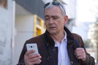 Şık, olgun bir adam sırt çantası ya da çantasıyla şehir kaldırımında yürüyor ve cep telefonunu kullanıyor.