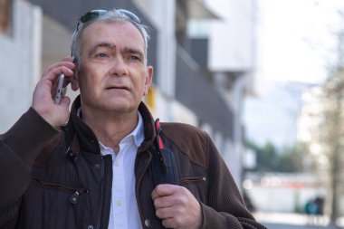 Şık, olgun bir adam sırt çantası ya da çantasıyla şehir kaldırımında yürüyor ve cep telefonunu kullanıyor.