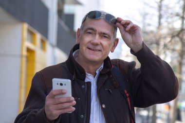 Şık, olgun bir adam sırt çantası ya da çantasıyla şehir kaldırımında yürüyor ve cep telefonunu kullanıyor.