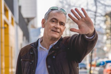 Şık, olgun bir adam kaldırımda yürüyor ve el sallıyor.