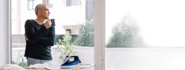 Adam evde gömleğini ütülerken kahve içip pencereden dışarı bakıyor.