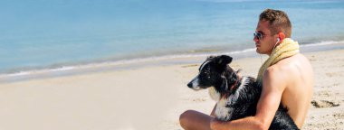 Genç bir adam köpeğiyle sahilde kulaklıkla müzik dinliyor.