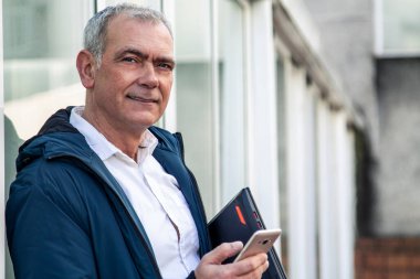 Yetişkin çekici bir adam cep telefonu kullanıyor ve dizüstü bilgisayarını sokakta tutuyor.