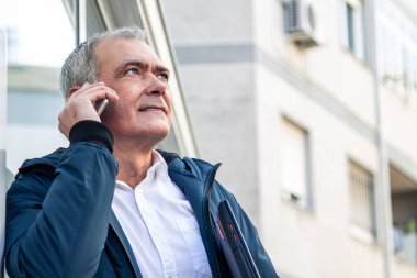 Yetişkin çekici bir adam cep telefonu kullanıyor ve dizüstü bilgisayarını sokakta tutuyor.