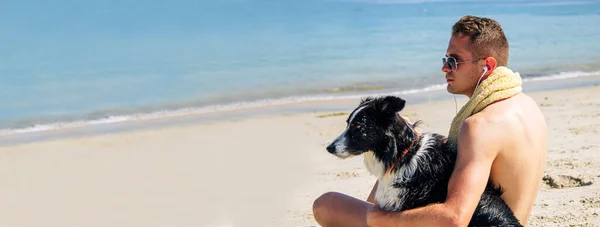 若い男が犬とビーチでヘッドフォンで音楽を聴き — ストック写真
