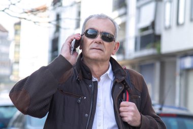 Şık, olgun bir adam sırt çantası ya da çantasıyla şehir kaldırımında yürüyor ve cep telefonunu kullanıyor.