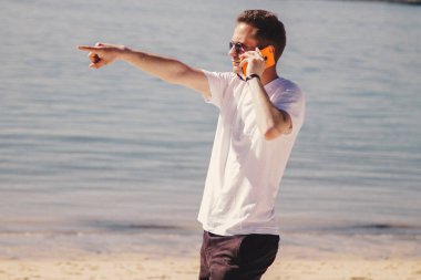 Genç adam kumsalda yürüyor cep telefonuyla konuşuyor ve parmağıyla işaret ediyor.