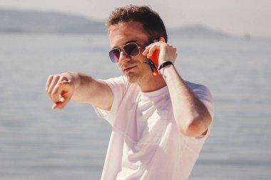 Genç adam kumsalda yürüyor cep telefonuyla konuşuyor ve parmağıyla işaret ediyor.