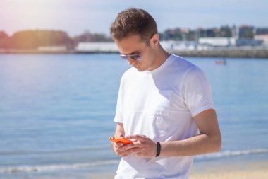 Gülümseyen genç adam rahat ve plajda cep telefonu kullanarak eğleniyor.