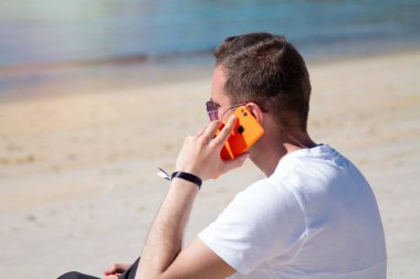 Gülümseyen genç adam rahat ve plajda cep telefonu kullanarak eğleniyor.