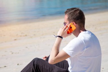 Gülümseyen genç adam rahat ve plajda cep telefonu kullanarak eğleniyor.