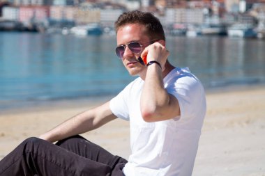 Gülümseyen genç adam rahat ve plajda cep telefonu kullanarak eğleniyor.