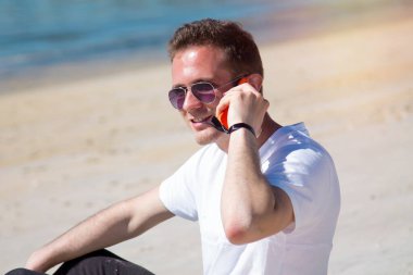 Gülümseyen genç adam rahat ve plajda cep telefonu kullanarak eğleniyor.