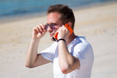 Gülümseyen genç adam rahat ve plajda cep telefonu kullanarak eğleniyor.